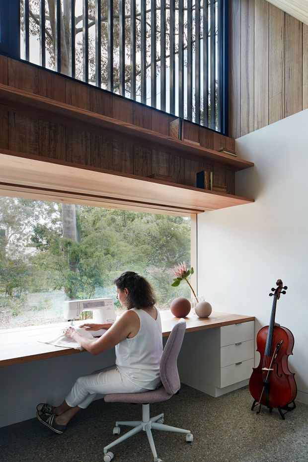 A bright, double-height home office or creative workspace with a built-in desk positioned beside large windows overlooking trees. The room features extensive dark timber wall panelling and shelving, a pale timber work surface, white built-in drawers, polished concrete-style flooring, and a high bank of vertical glazed openings with dark frames. A person is sewing at the desk, with a cello resting against the wall and small decorative objects on the worktop.