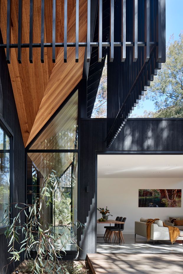 A dramatic modern residential interior/exterior threshold with a double-height glazed living area. The composition highlights a dark black-clad structure, exposed timber-lined soffit, deep vertical metal fins, tall windows, and an open-plan living and dining room visible beyond. Strong daylight filters through surrounding trees, creating patterned shadows across the floor and walls.