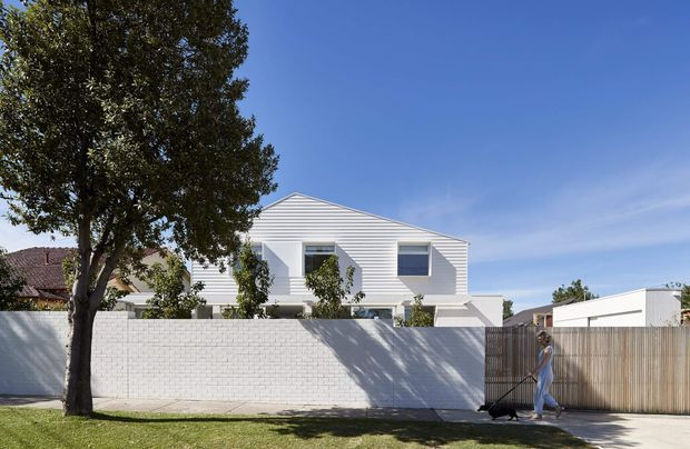 Street-facing exterior view of a contemporary white-clad residential building behind a low white masonry boundary wall. The two-storey facade has horizontal cladding, deep-set rectangular windows, projecting geometric forms, and a flat or very low-pitched roofline. A timber-slatted gate and driveway sit to the right, with mature landscaping and neighbouring homes in the background.