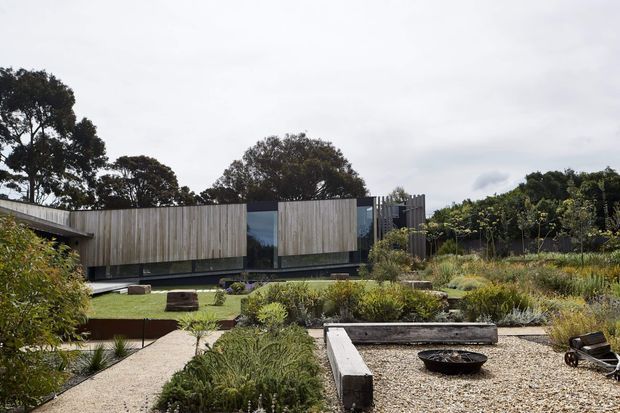 A low, contemporary residential building set within a landscaped garden. The facade combines extensive vertical timber cladding with dark framed glazing and darker vertical screening elements. A paved path leads through layered ornamental planting toward the house, with a gravel outdoor area and a built-in fire bowl in the foreground. The image is an exterior architectural and landscape view under overcast light.