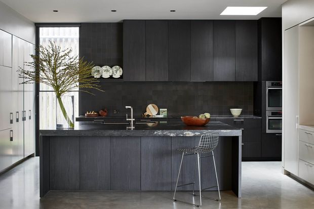 A sophisticated contemporary kitchen with a large central island, dark vertical-grain cabinetry, dark stone benchtops, and a dark tiled splashback. White floor-to-ceiling cabinetry lines the left side, while integrated appliances are concealed in the rear joinery. A tall window brings in natural light, complemented by recessed ceiling lights and a large square skylight. The space has a monochromatic, refined appearance with a wire-frame bar stool and minimal decorative styling.