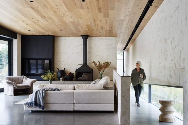 Open-plan contemporary living room with a large light fabric sectional sofa, sculptural lounge chairs, and a central freestanding black wood-burning fireplace. The room features a warm timber-clad ceiling, polished grey concrete flooring, dark built-in cabinetry, expansive glazing, and a textured pale feature wall. A partially visible lower-level circulation or stair area appears beyond the half-height wall.