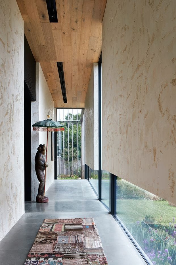 A narrow contemporary hallway or gallery corridor with full-height glazing along one side, textured plaster walls, a warm timber-lined ceiling with recessed linear lighting, and a polished grey concrete floor. A patterned runner and decorative statue beneath a fringed parasol add eclectic character. The glazing overlooks planted landscaping.