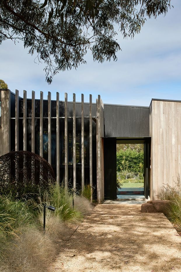 Exterior view of a contemporary single-storey building set within landscaped grounds. The facade combines dark vertically ribbed cladding, pale timber-like vertical screening and a recessed entrance passage. A gravel path leads through naturalistic planting toward the doorway, with a garden and pool-like blue surface visible beyond. Overhanging eucalyptus branches frame the scene.