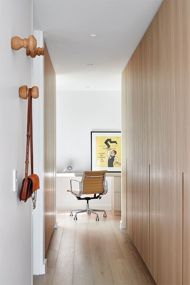 A narrow, bright home office viewed through a timber-panelled hallway. The space features warm light timber flooring and full-height vertical timber wall cabinetry or panelling, white walls and ceiling with recessed downlights, a compact white desk, and a mid-century style tan leather office chair. A framed vintage poster provides a small visual accent. Timber-handled accessories are mounted on the left wall.