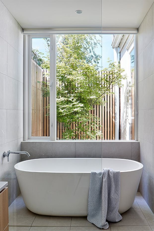 Bright contemporary bathroom featuring a freestanding white bathtub positioned beneath a large window. The room has pale grey tiled walls and floor, a simple wall-mounted bath tap, a roller blind, and a planted courtyard with timber screening visible through the glazing. A large mirror reflects the window and bathroom interior.