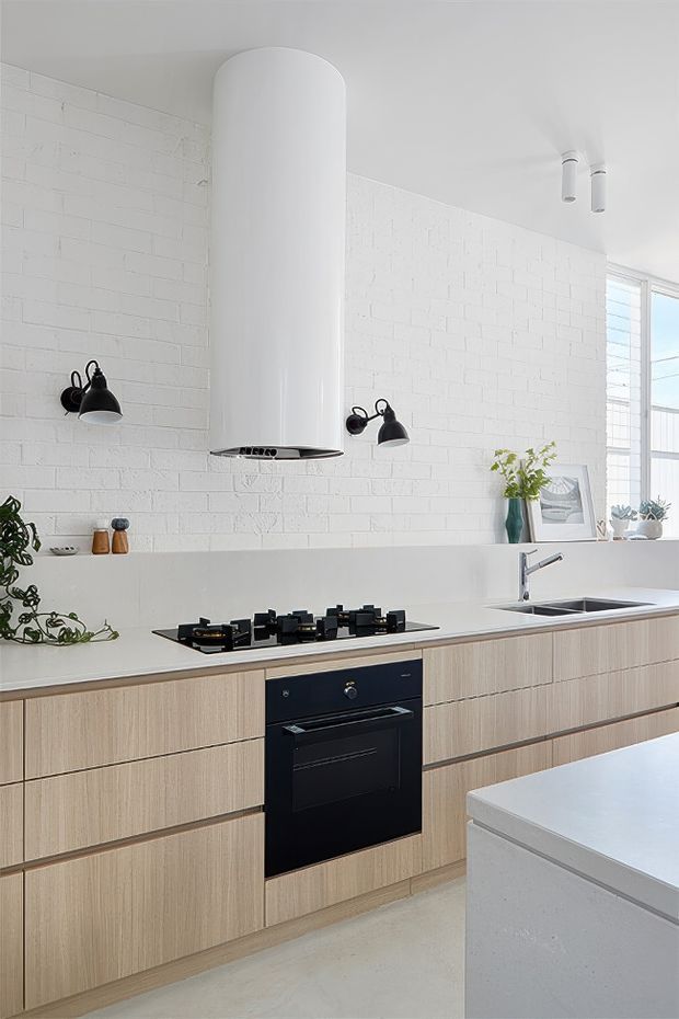 Bright contemporary kitchen with a long run of light timber-grain cabinetry, white benchtops, an integrated black oven and gas cooktop, and a large white island in the foreground. The kitchen features a prominent cylindrical white rangehood, white-painted exposed brick walls, black wall sconces, stainless-steel tapware and sink, and ample natural light from louvred glazing.