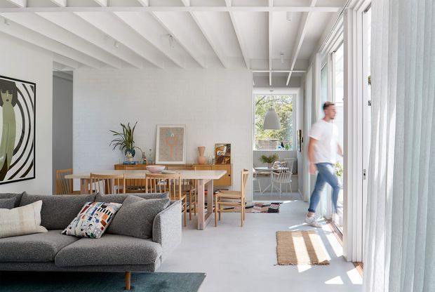 Bright open-plan living and dining area with a contemporary white interior, exposed painted ceiling beams, full-height glazed doors, a grey sectional sofa, timber dining table and chairs, and abundant natural light. A small outdoor or glazed sitting area is visible beyond the dining space.