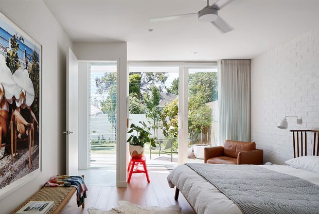 Bright contemporary bedroom with a large upholstered bed, white-painted brick feature wall, white ceiling, pale timber flooring, and expansive glazed doors opening to a garden. A leather lounge chair, bedside lighting, indoor plants, artwork, bench seating, and a ceiling fan furnish the room. The space has an airy, relaxed feel with strong indoor-outdoor connection.