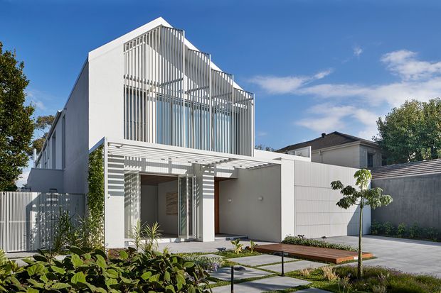 Modern white multi-storey residence viewed from the landscaped front garden. The facade features strong geometric forms, extensive floor-to-ceiling glazing screened by vertical white fins, a projecting pergola over the entry, and a paved driveway or forecourt. Low planting, stepping stones, timber decking, and high boundary walls complete the contemporary streetscape.