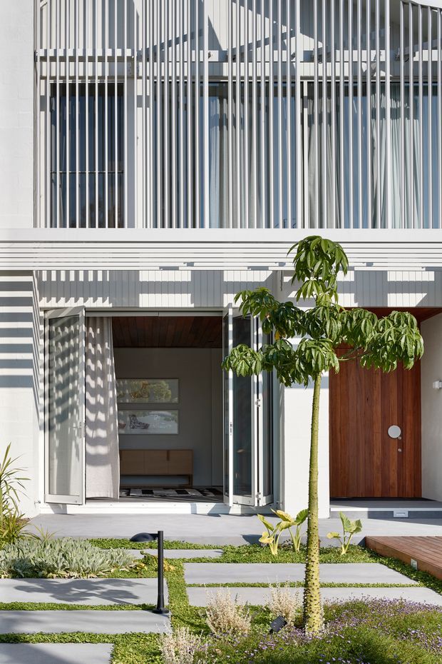 Bright contemporary multi-level residential facade viewed from the landscaped front garden. The white facade is screened by dense vertical fins and horizontal bands, with extensive glazing, a tall timber pivot door, and a recessed entry opening. A structured garden path of large concrete pavers, low groundcover, planting, and a small timber deck leads to the house.