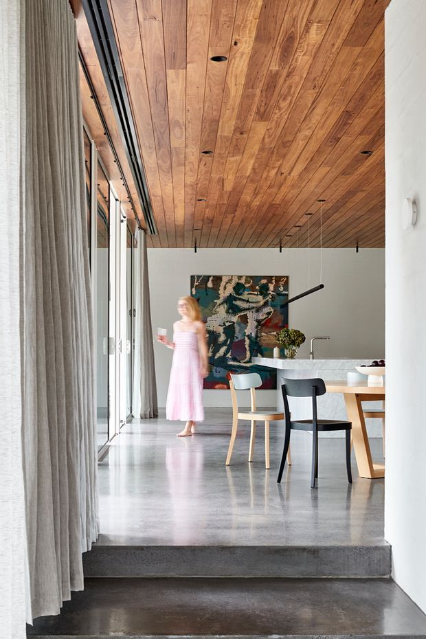 A refined open-plan dining and kitchen interior viewed from a lower level, with a polished concrete floor, full-height glazed wall concealed by sheer curtains, a warm timber-lined ceiling with recessed downlights, white walls, and minimalist black pendant lighting. A dining table and sculptural chairs sit beside a pale stone kitchen island, while a large colourful abstract artwork provides a focal point. A short concrete step separates the foreground from the main living area.