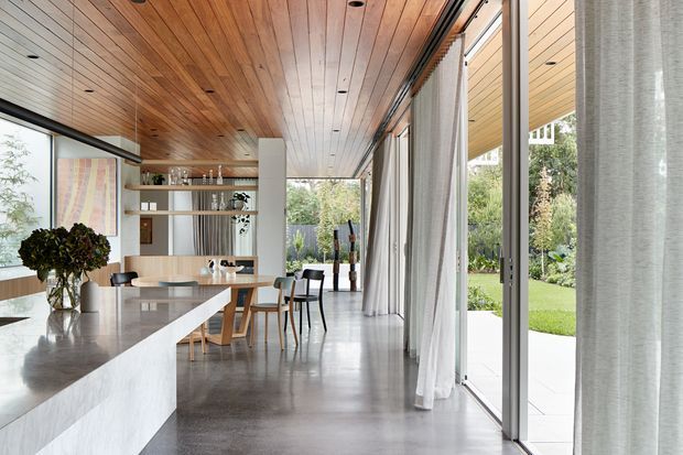 Bright open-plan contemporary interior showing a kitchen and dining area. A long pale stone island dominates the foreground, with a timber dining table and chairs beyond. Full-height glazed sliding doors with sheer curtains open to a landscaped garden. The room features extensive warm timber ceiling lining, polished grey concrete flooring, white built-in shelving, and minimalist integrated lighting.