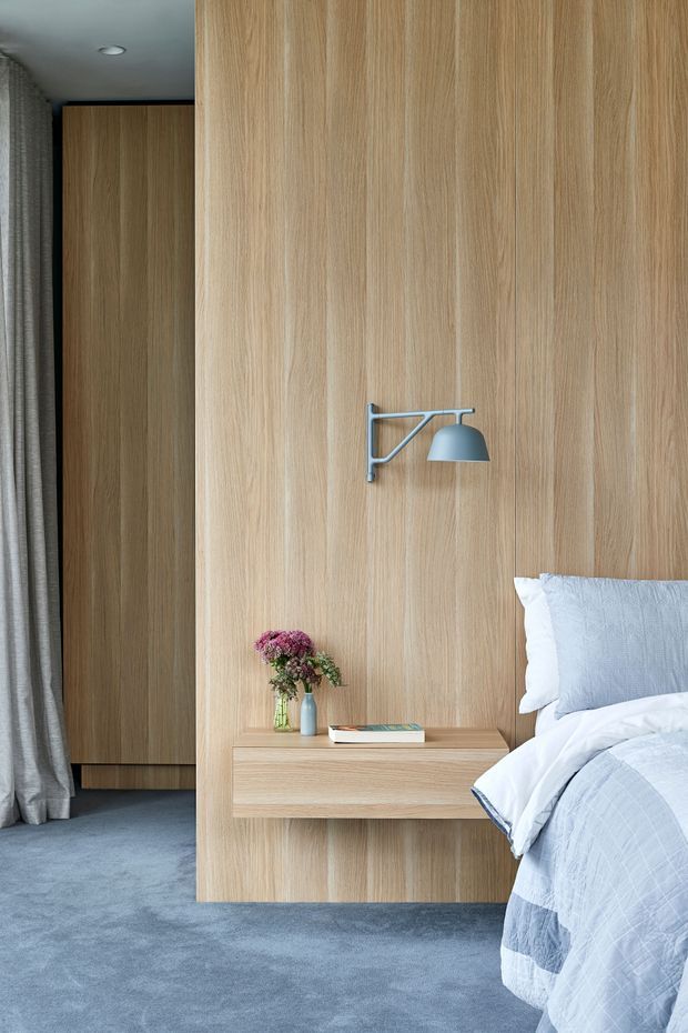 A refined bedroom interior featuring a full-height light timber wall and matching floating bedside table, a minimalist wall-mounted metal reading lamp, soft blue-grey carpet, and a partially visible bed dressed in white and pale blue bedding. Floor-to-ceiling curtains and integrated timber joinery are visible at the left.