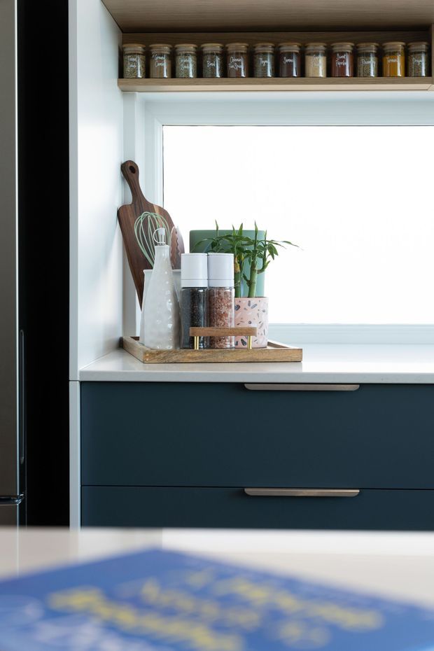 Close-up view of a refined kitchen storage area beside a bright window. Deep navy-blue drawers with brushed metal pulls sit beneath a white countertop and white-framed window. An open timber shelf above holds labelled spice jars, while a timber tray displays a cutting board, whisk, bottles, canisters, and a small potted plant. The scene has a clean, contemporary feel with warm timber accents.