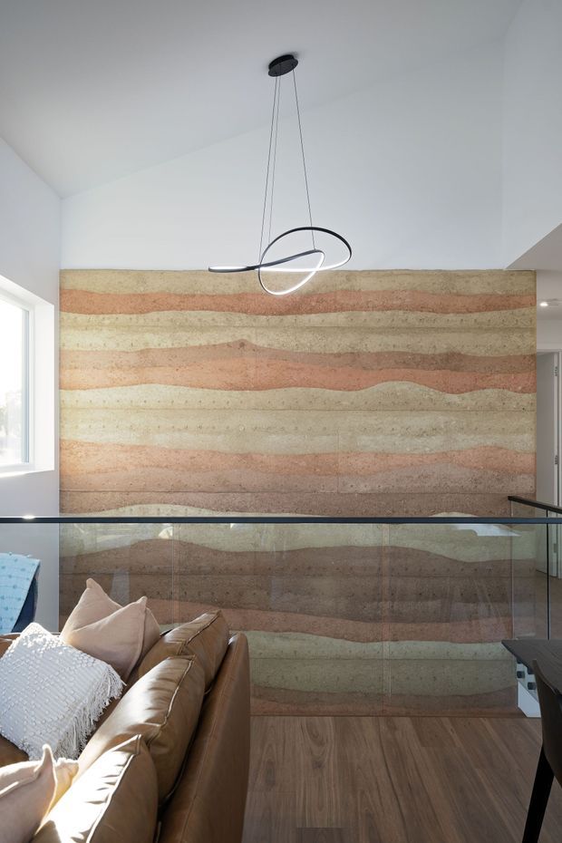 A bright contemporary living area viewed from an upper level, featuring a dramatic layered earthen feature wall in cream, terracotta and muted pink tones. A glass balustrade overlooks the room, with a tan leather sofa in the foreground and a sculptural looping pendant light suspended from the white ceiling. Timber flooring and abundant daylight complete the space.