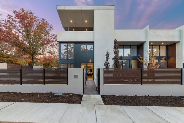 Street-facing view of a contemporary multi-level residence with a strong geometric facade, white rendered walls, dark vertical cladding, large glazed openings, recessed soffit lighting, and a dark horizontal-slatted privacy fence. A concrete path leads through the front gate to the entry, with landscaped garden beds and autumn trees framing the property.