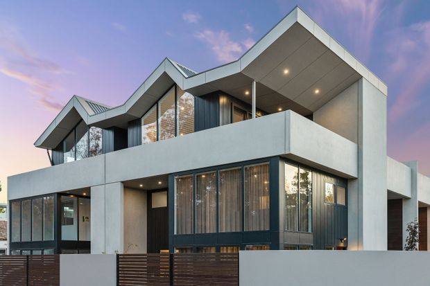 Contemporary multi-storey residential house viewed from the street at dusk. The angular facade features projecting upper-level balconies and deep roof overhangs, large glazed openings with dark frames, pale rendered or concrete-like wall surfaces, and horizontal timber screening along the front boundary.