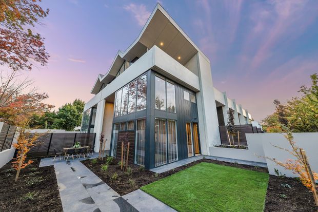 Exterior view of a contemporary multi-storey residence with a sharply folded gable-like roof form, angular concrete walls, extensive floor-to-ceiling glazing, and dark framed windows. The home is set within a landscaped suburban garden with paved stepping paths, lawn, raised planting beds, boundary fencing, and a small outdoor dining area. The image is photographed at dusk with warm interior lighting visible through the windows.