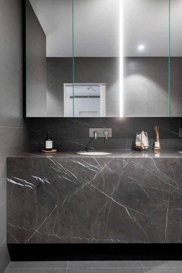 A contemporary bathroom vanity with a large frameless mirrored cabinet, dark grey marble-look wall and vanity surfaces with prominent white veining, an integrated white basin, and minimalist wall-mounted tapware. Black floor tiles and restrained accessories create a dark, polished, luxurious appearance.