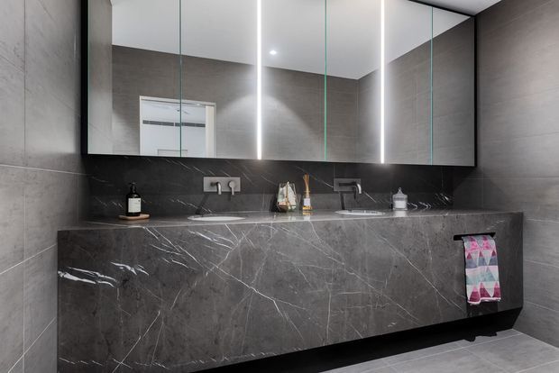 Contemporary bathroom vanity with a long double-basin stone counter and integrated cabinetry, set against dark grey large-format wall tiles. A mirrored wall cabinet with vertical LED lighting spans the vanity, creating a sleek, high-contrast, luxurious finish.