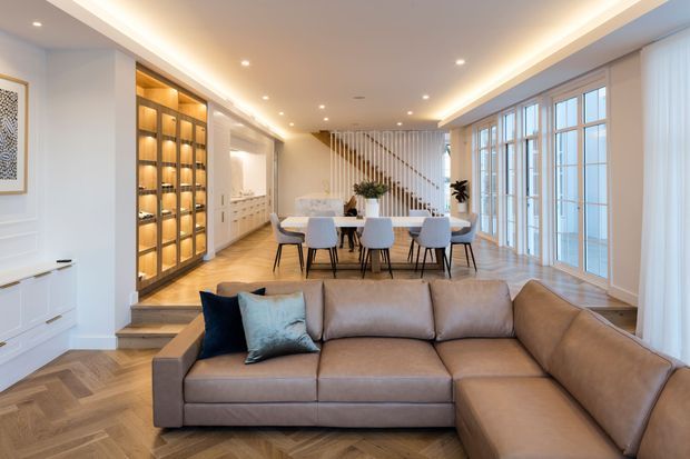 A bright, refined open-plan interior combining a living room, dining area and kitchen zone. A large taupe L-shaped sectional sits in the foreground on herringbone timber flooring, while a dining table with upholstered chairs occupies the centre. Floor-to-ceiling glazed doors and windows line the right side, and illuminated built-in display cabinetry, white joinery and a sculptural slatted stair screen add architectural detail. Recessed and concealed lighting create a warm, inviting atmosphere.