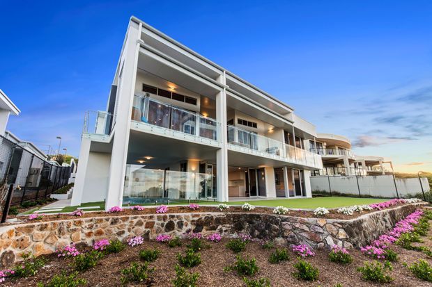 Exterior view of a contemporary multi-level residential development with white rendered walls, extensive glass balcony balustrades, recessed outdoor areas, and large glazed openings. The building sits above landscaped terraced stone retaining walls with flowering groundcover, and neighboring residential units are visible at the sides.