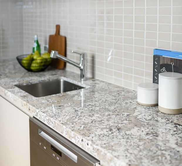 Close-up view of a contemporary kitchen worktop featuring a mottled grey and white stone benchtop, undermount stainless-steel sink, chrome mixer tap, pale square-tile splashback, and integrated stainless-steel dishwasher. A fruit bowl, chopping board, and ceramic canisters sit on the counter.