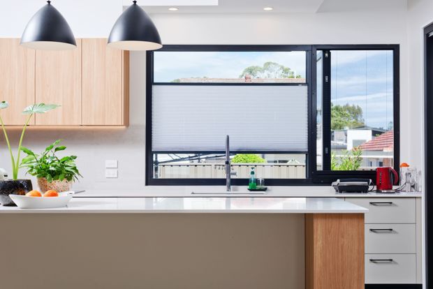 Bright contemporary kitchen with a large island, pale timber overhead and island cabinetry, white benchtops, dark-framed windows, and matte black pendant lights. The room overlooks a suburban setting and includes a sink beneath the window, integrated storage, and modern appliances.