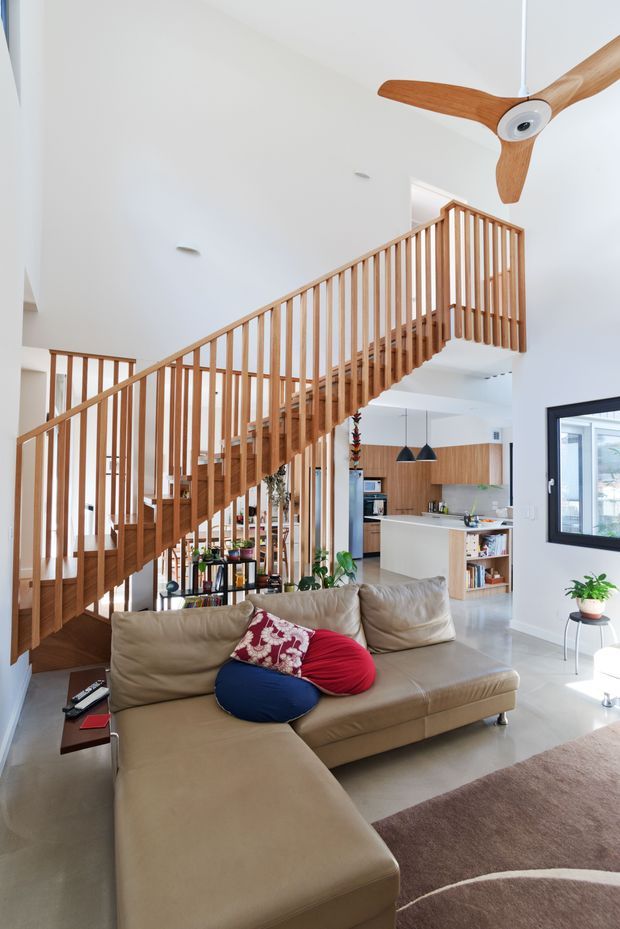 Bright open-plan living room with a large beige sectional sofa in the foreground, a dramatic timber staircase with vertical balustrades, and a modern kitchen visible behind it. White walls and a high ceiling create an airy interior, with timber cabinetry, pendant lights, indoor plants, and a sculptural timber ceiling fan.