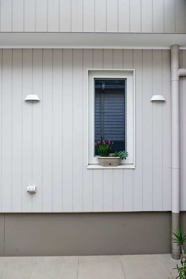 Close-up exterior view of a residential wall clad in pale grey vertical boards, with a small white-trimmed window, exterior wall lights, exposed downpipe, and potted plants on a paved surface. The composition is simple and contemporary with a restrained neutral palette.