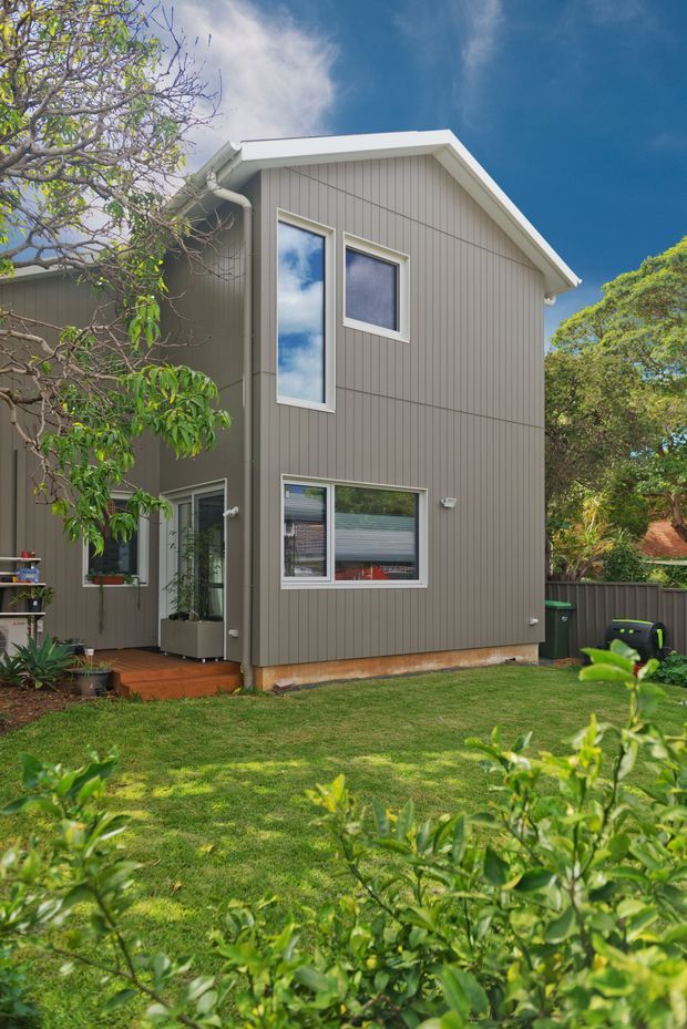 Exterior view of a compact two-storey residential building with a simple gabled roof, grey vertical weatherboard cladding, white window and roof trims, and several vertically oriented windows. The house sits beside a timber deck and lawn within a leafy suburban garden, with fencing and outdoor bins visible at the side.