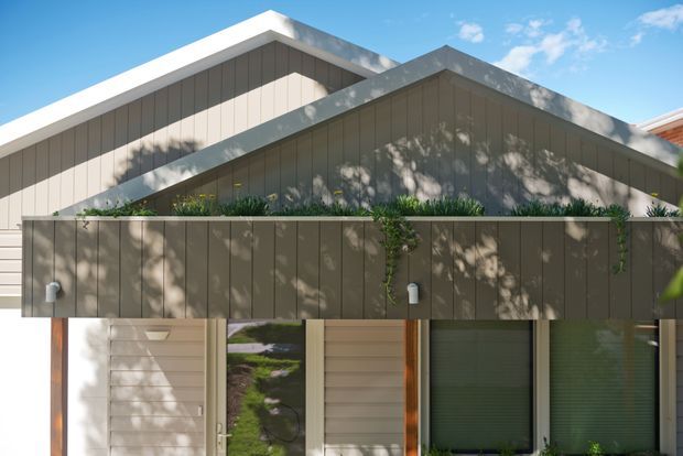 Exterior view of a contemporary low-rise residence with overlapping pitched gable roof forms, vertical dark grey cladding, and a projecting upper planter or balcony structure. Ground-level glazing and a timber-toned entry surround sit beneath the sheltered upper volume. Strong sunlight casts tree shadows across the facade against a clear blue sky.
