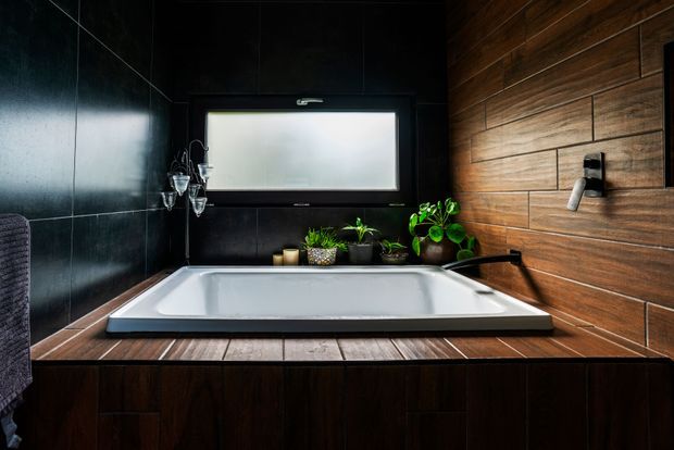Dark, moody bathroom featuring a built-in white bathtub set into a dark timber-clad surround. The walls combine glossy black panels on the left with horizontal warm wood panelling on the right, including a wall-mounted bath filler. A frosted rectangular window, potted plants, and candle holders add atmospheric detail.