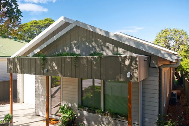 Exterior view of a compact contemporary gable-roofed house or extension. The facade combines dark vertical timber-look cladding, pale horizontal weatherboard siding, large glazed openings with timber-toned frames, and a covered front verandah or entry canopy. Lush residential landscaping surrounds the building, with strong sunlight and tree shadows across the facade.
