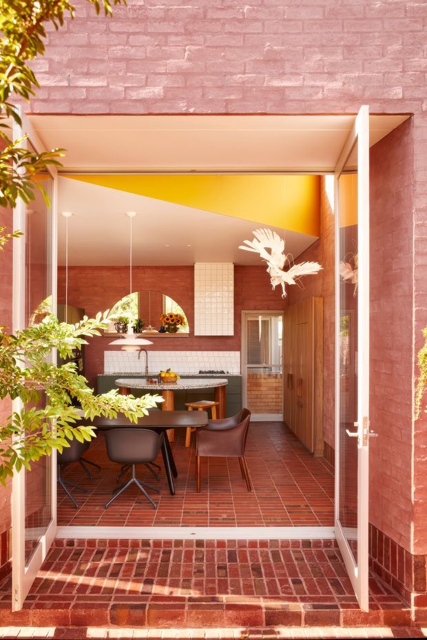 A bright architectural view through open glass doors into a compact brick-lined kitchen and dining area. The space features red brick walls and tiled flooring, white tiled splashbacks, warm timber cabinetry, a dark stone island benchtop, and a vivid yellow ceiling or upper wall plane. A small dining table with modern dark chairs sits in the foreground, while plants and a sculptural hanging light add decorative detail. The image is photographed from an exterior threshold looking inward.