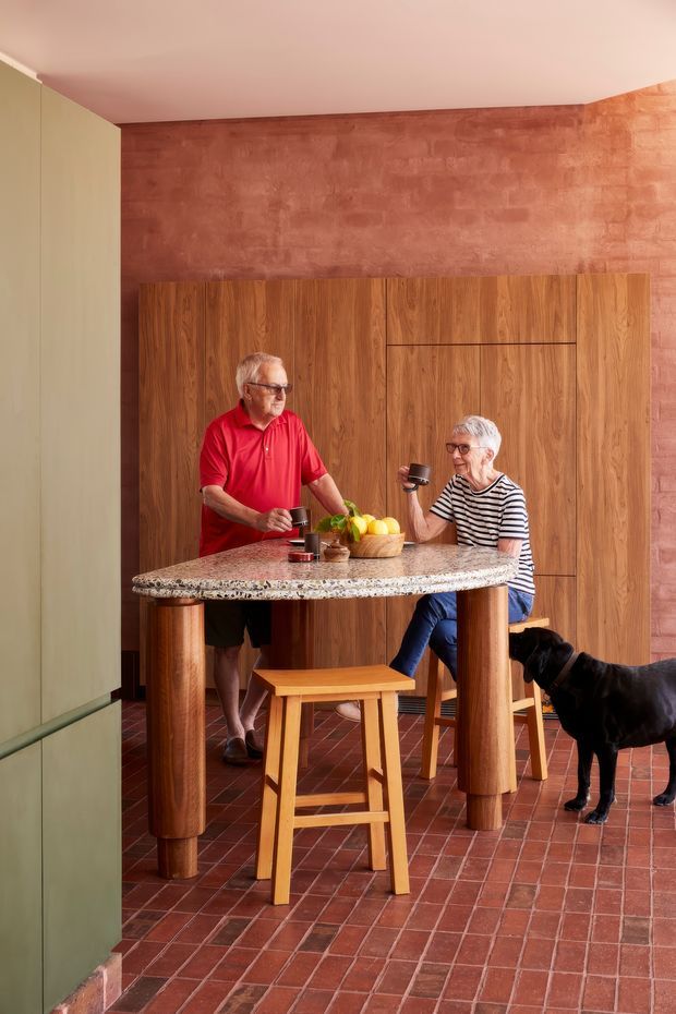 A warm, richly textured kitchen interior featuring a tall island with a speckled stone benchtop and cylindrical timber supports. Full-height timber cabinetry spans the back wall, paired with a terracotta-toned textured wall and reddish brick floor tiles. Two people are gathered at the island with drinks, while a black dog stands nearby. A muted green cabinet is visible at the left edge.