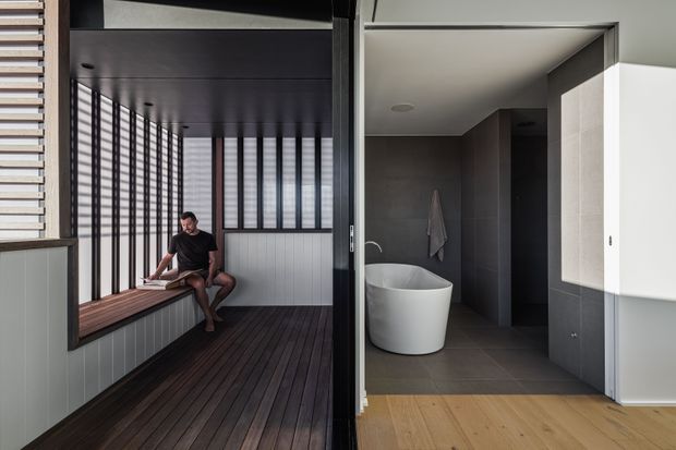 A contemporary interior showing a dark timber-lined enclosed balcony or sunroom with a built-in bench and vertical screening beside a minimalist bathroom. The bathroom features dark large-format wall and floor tiles, a white freestanding oval bathtub, a wall-mounted towel, and a pale stone or tile vanity surface. Timber flooring leads toward the bathroom, while strong daylight enters through the screened glazing.