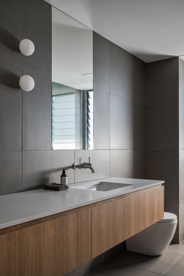 A refined contemporary bathroom with a floating timber-veneer vanity, integrated white basin, wall-mounted chrome tapware, and a partially visible white toilet. Large-format dark grey wall and floor tiles create a sleek, moody backdrop, while a full-height mirror reflects a window with horizontal blinds. Minimal spherical wall lights and restrained styling complete the space.