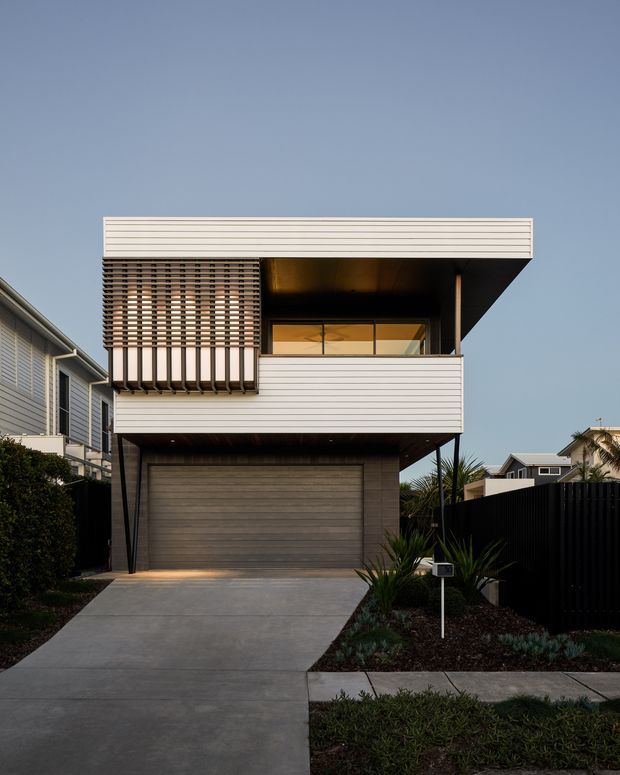 Street-facing exterior view of a contemporary multi-level residence with a projecting upper floor, flat roof, horizontal light-coloured cladding, dark timber screening, recessed balcony or window bay, and an integrated double garage. A concrete driveway and restrained low-maintenance planting lead to the entrance, with neighbouring homes visible on either side.