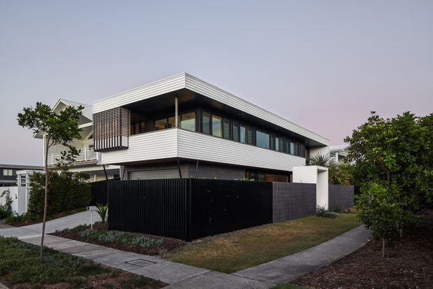 Contemporary two-storey residential house viewed from the street at dusk. The home has a strong horizontal composition, a low-pitched or flat roofline, white horizontally lined cladding, dark recessed glazing and framing, a projecting upper-level balcony, and a tall black slatted boundary fence. Paved walkways, lawn, and established garden planting surround the property.