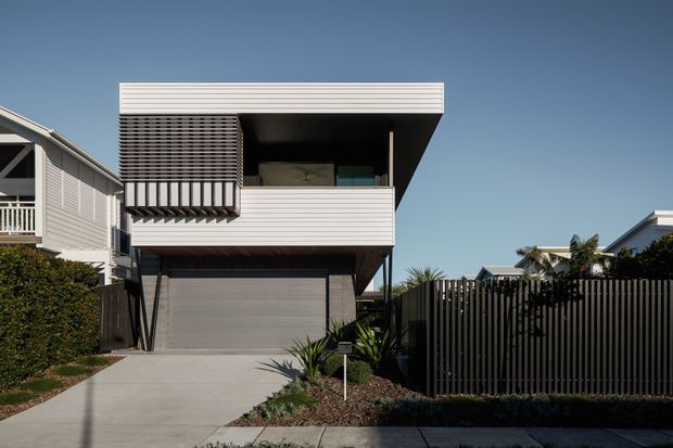 Front exterior view of a contemporary multi-level residence with horizontal light-coloured cladding, a projecting upper-floor balcony, dark screened balcony enclosure, integrated ground-level garage, and a flat roof. A concrete driveway, vertical privacy fencing, and minimal landscaped planting define the frontage.