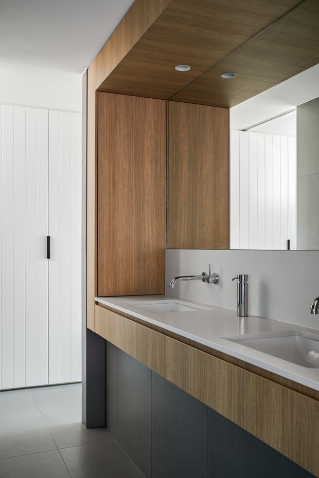 A contemporary bathroom vanity with a long double-sink counter, integrated white basins, chrome wall-mounted and deck-mounted tapware, and a large mirror. Warm timber-grain cabinetry extends across the vanity, tall storage, and ceiling, contrasted with dark lower joinery, pale grey porcelain floor tiles, and white vertically panelled walls. The space is clean-lined and softly lit, with a partially visible adjoining area beyond the vanity.