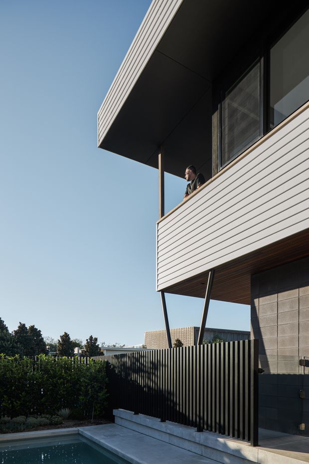 Photograph of a contemporary multi-storey residential exterior with white horizontal cladding, a deep projecting upper-level balcony, dark metal framing, and angled structural supports. A black vertical-slat fence, landscaped garden, paved terrace, and the edge of a swimming pool are visible at ground level. The image is taken from outside under a clear sky, emphasizing the building facade and cantilevered form.