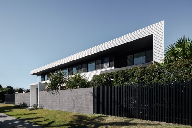 Exterior photograph of a contemporary two-storey residence with a strong horizontal form and deep projecting upper-level roof or fascia. The facade combines dark recessed glazing and cladding with crisp white horizontal siding, while a tall black vertical-slat fence, concrete boundary wall, lawn, and tropical planting define the front yard.