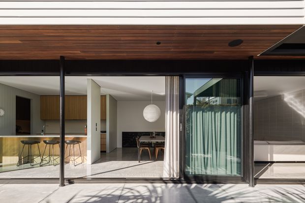 Wide view from an exterior terrace into a contemporary single-level residence through full-height glazed sliding doors. The composition shows a warm timber-lined soffit, slim dark window and door frames, an open-plan kitchen and dining area, and a tiled bathroom with a freestanding bathtub. Strong indoor-outdoor connection and clean modern detailing dominate the image.