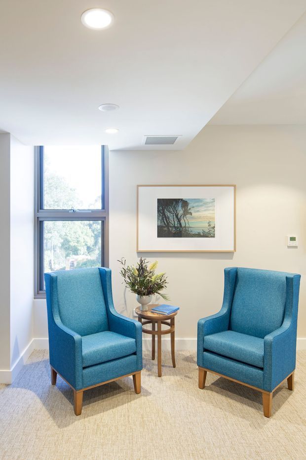 Bright, compact sitting area with two blue upholstered wingback armchairs arranged around a small round timber side table. The room has light carpet, off-white walls and ceiling, a tall window with timber-toned framing, recessed ceiling lights, and a framed landscape artwork.