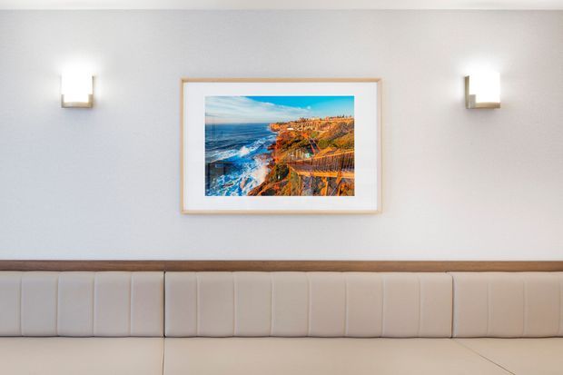 A minimalist interior wall featuring a long upholstered cream bench or banquette with vertical channel tufting and a timber trim rail. Two modern wall sconces flank a framed coastal landscape photograph. The palette is light and neutral with warm timber accents.