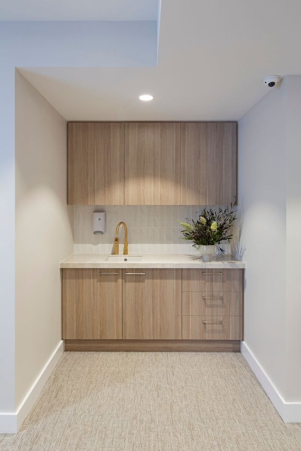 A compact, neatly fitted kitchenette or utility kitchenette with light wood-grain upper and lower cabinetry, a pale solid-surface benchtop, small undermount sink, and brass gooseneck tap. The cabinetry is set against a light tiled splashback and pale painted walls, with beige carpet flooring and recessed ceiling lighting. A flower arrangement and wall-mounted dispenser sit on the benchtop, and a security camera is visible near the ceiling.