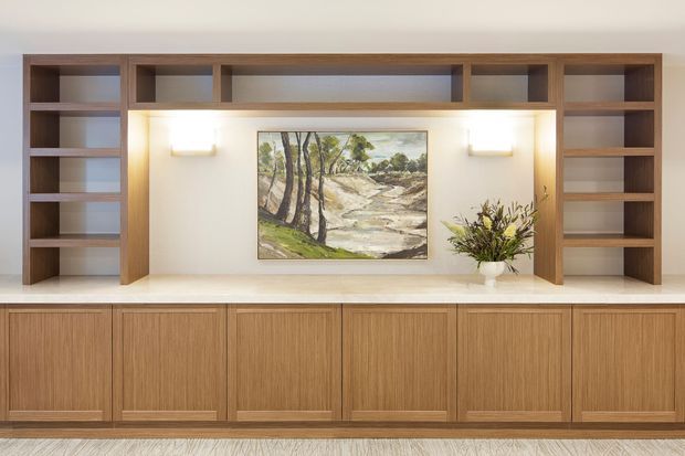 A built-in timber wall unit spans the width of the room, combining open shelving, vertical display niches, and lower cabinetry with integrated horizontal pulls. The central recessed niche has warm wall lighting, a landscape artwork, and a small vase arrangement. The joinery is set against light painted walls and a pale stone-like countertop, with a neutral textured floor visible below. The composition is clean, symmetrical, and understated.